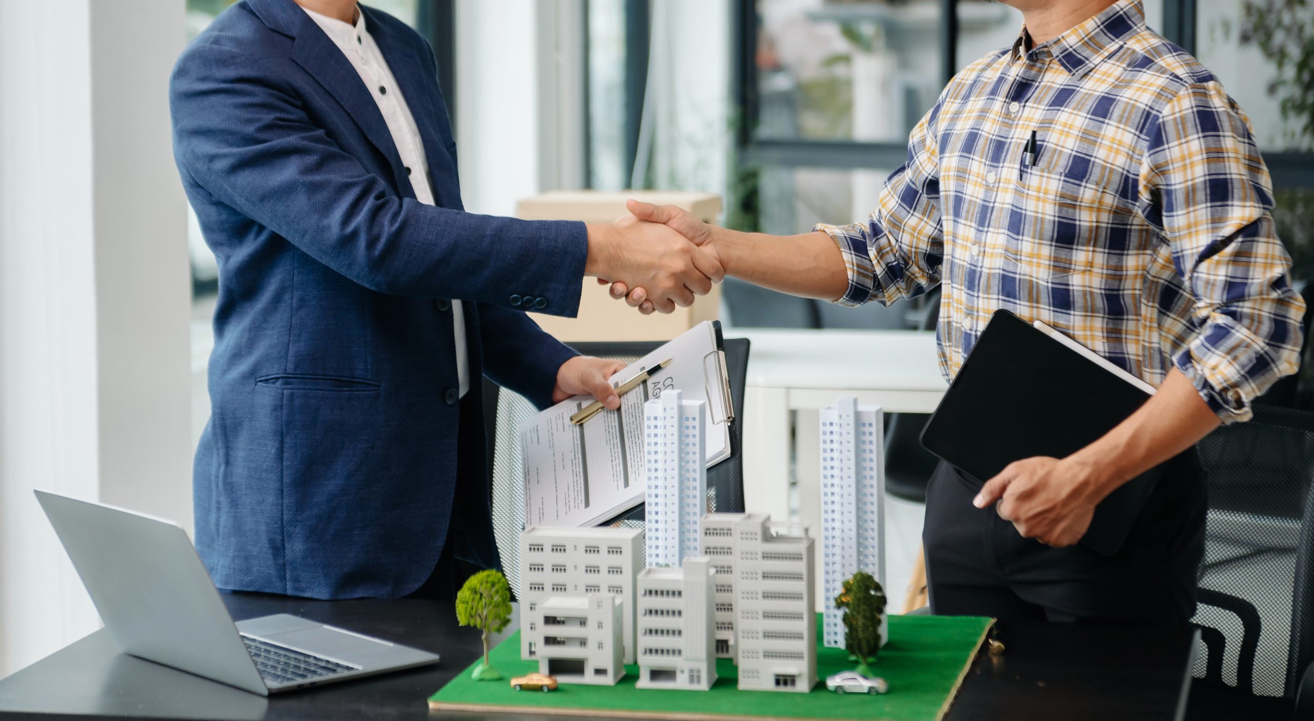 Handshake between two professionals over real estate property models during a commercial transaction.