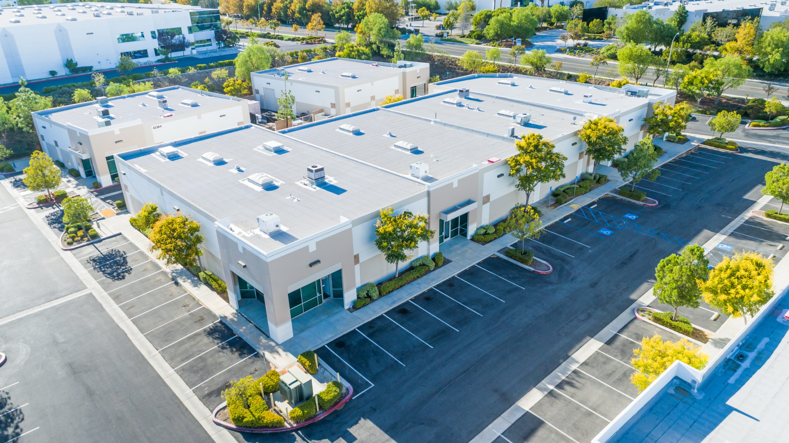 Aerial view of low-rise commercial buildings surrounded by parking areas, trees and nearby roads.
