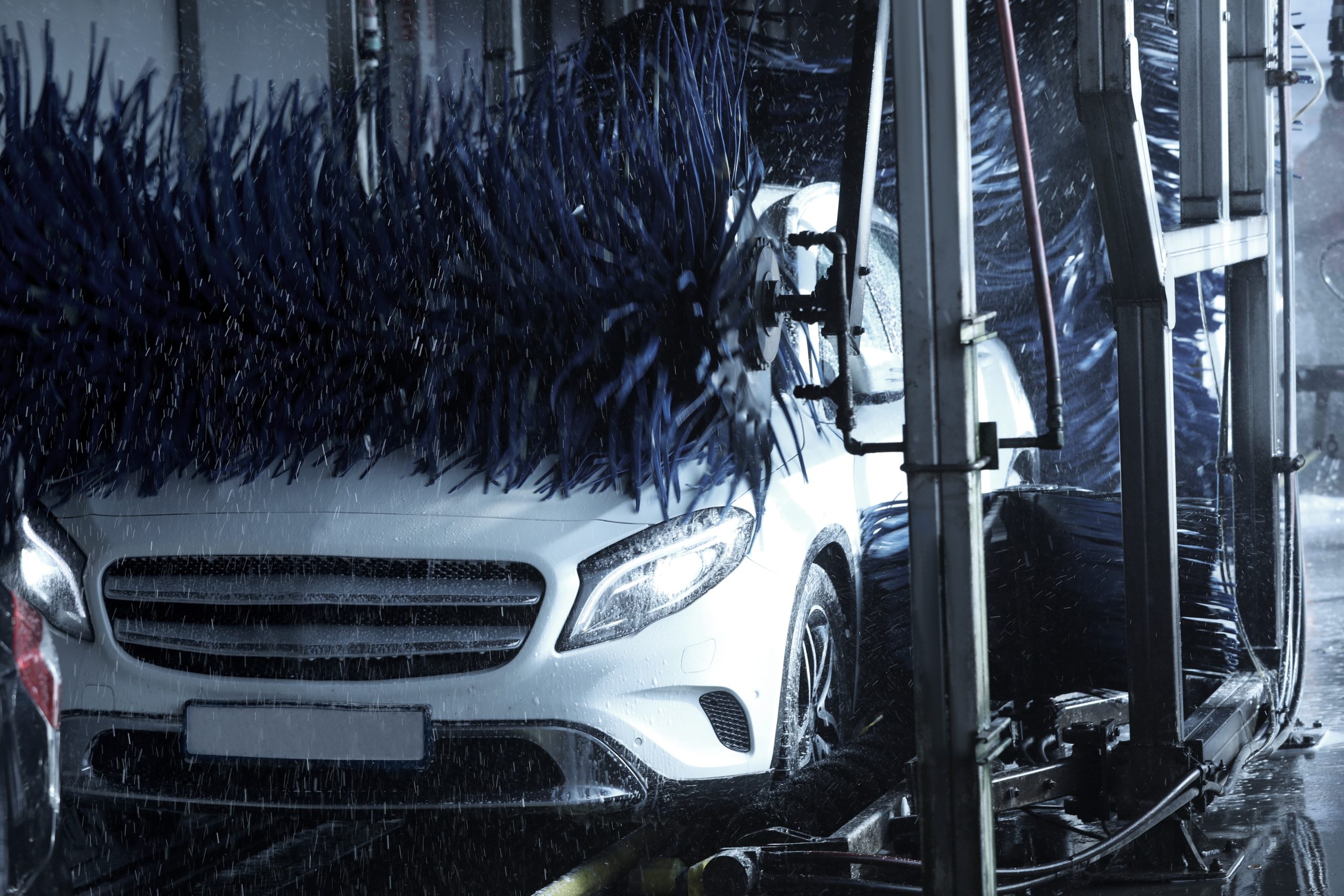White vehicle moving through an automatic car wash with rotating brushes and water spraying across its exterior.